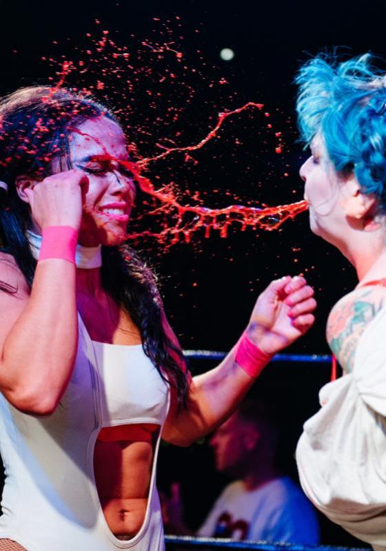 A performer with bright blue hair darts red liquid out of their mouth at their opponent hitting them square in the face