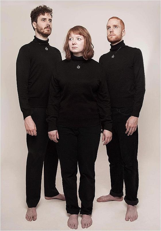 The 3 members of tarot dressed in all black, standing & looking off into the distance.