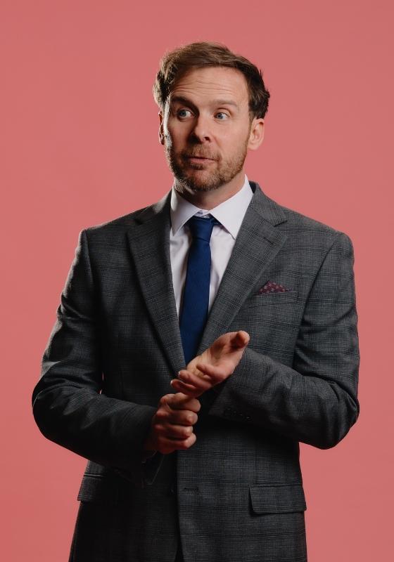 Colin standing in a check suit gesturing with his hands.