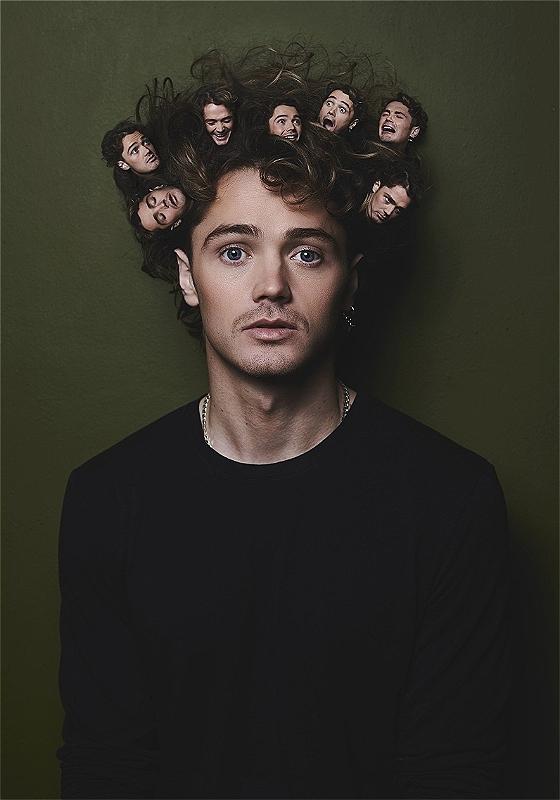 The performer looks into the camera in front of a dark green background. There are small pictures of his face in his hair.  