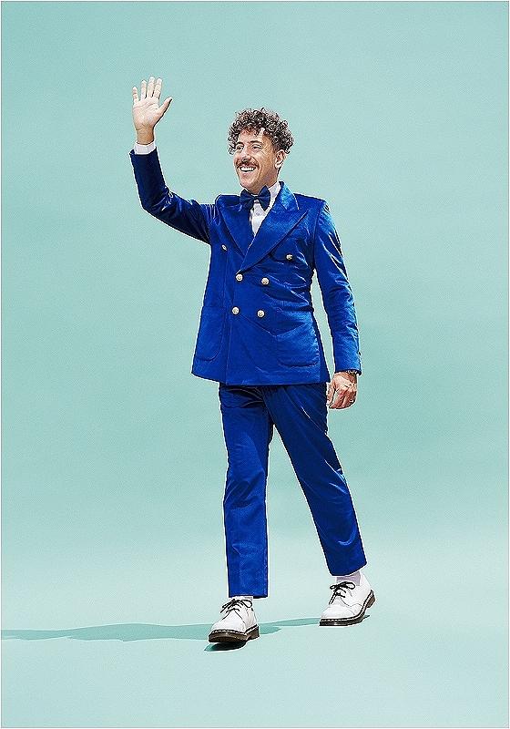 On a turquoise background, the performer is wearing a bright blue suit, smiling and waving their right hand. 