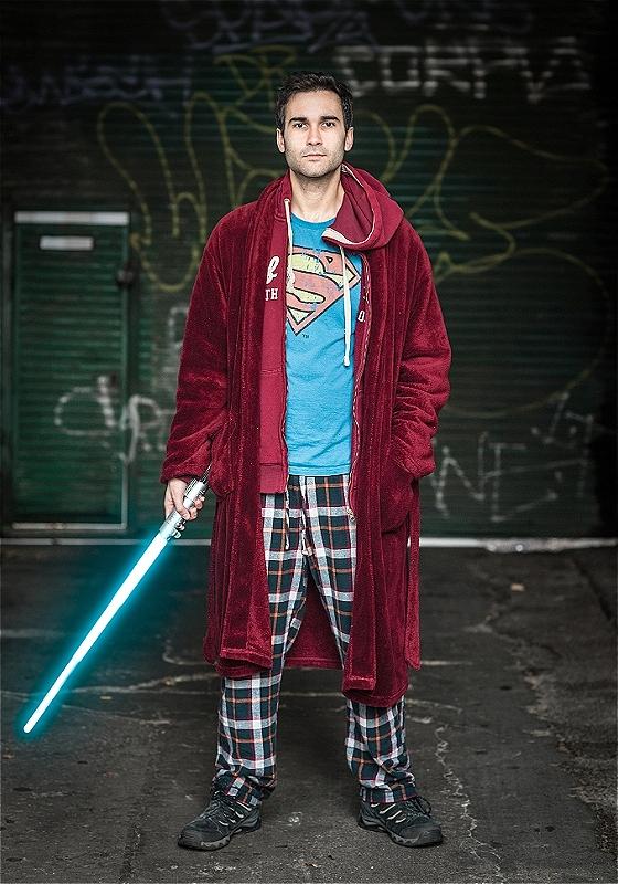 A performer stands facing the camera holding a lightsabre. They wear pyjamas and blue superman t-shirt. 