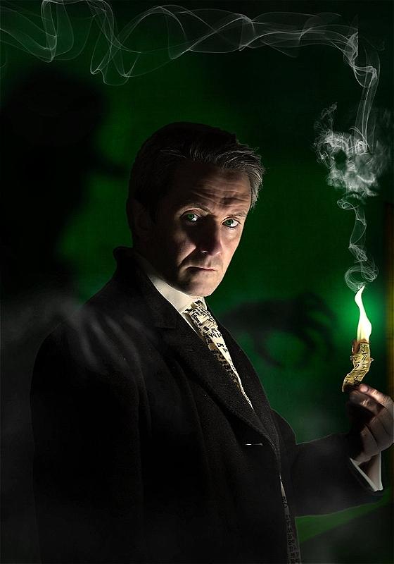 A dark background and a performer looking to the camera holding a candle with a green flame