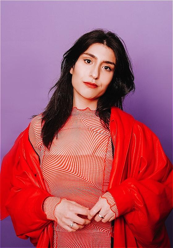 The performer wears a red dress and jacket in front of a purple background.