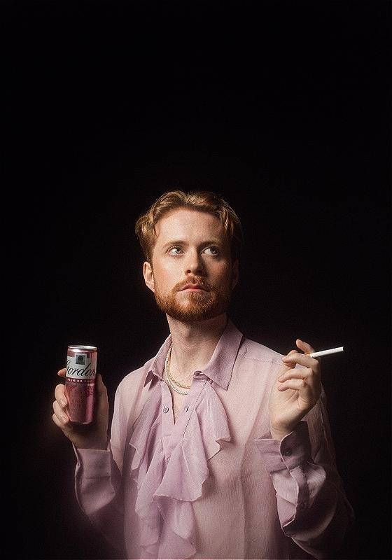 The performer wears a pink blouse and looks upwards. They hold a cigarette in one hand and a can of pink gin in the other. 