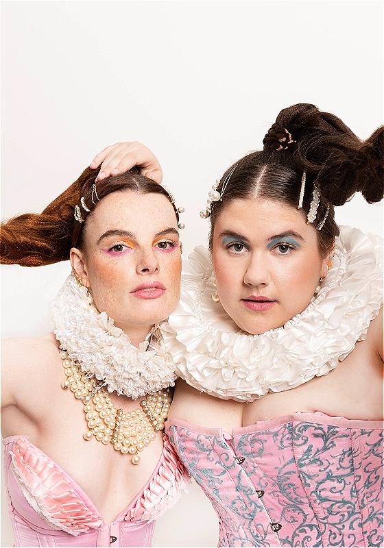 Two performers in  pink corsets and white collars. Both have ponytails and colourful eyeshadow.