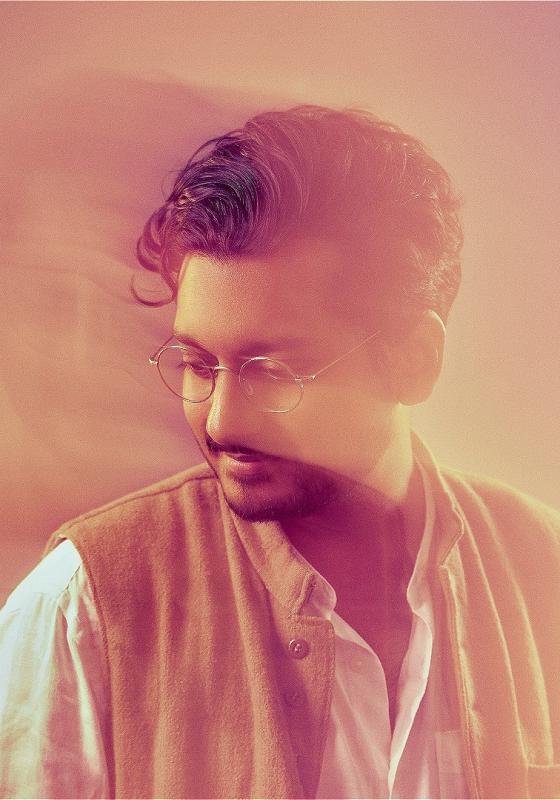 The performer looks to the bottom left, wearing glasses, a white shirt and beige sleeveless jacket. 