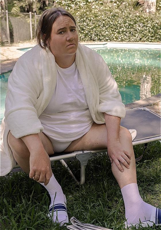 The performer sits in a garden in front of a swimming pool, wearing a white dressing gown.