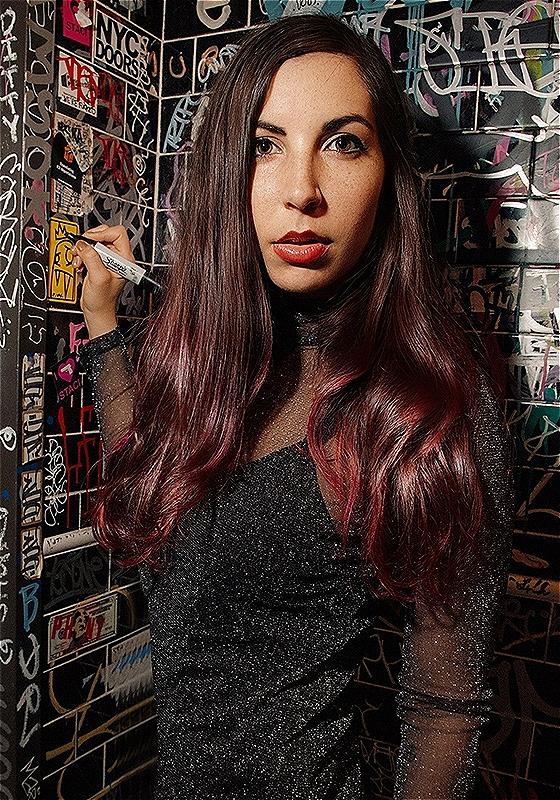 A close-up of the performer who wears a black glittery dress. They are writing on the wall behind them.