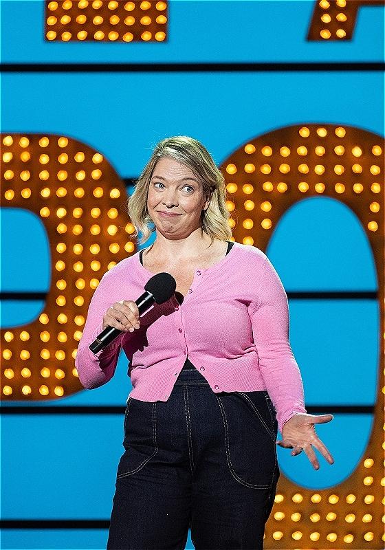 The performer is on stage at Live at the Apollo wearing a pink top and holding a microphone
