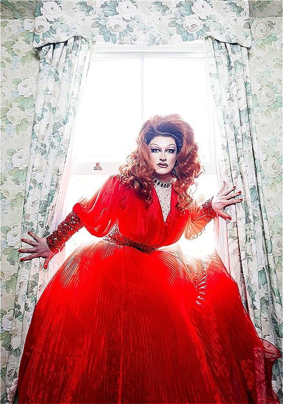 The performer is standing in front of an open window with floral curtains. They are wearing a red dress and look to the camera with a shocked expression.