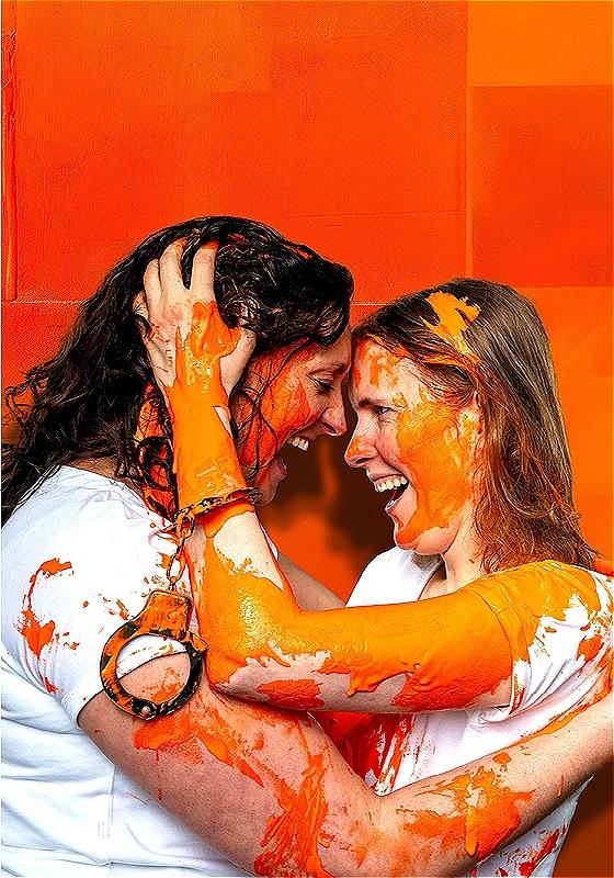 Two woman facing each other with a liberated expression. They are both covered in orange paint and one wears unchained handcuffs