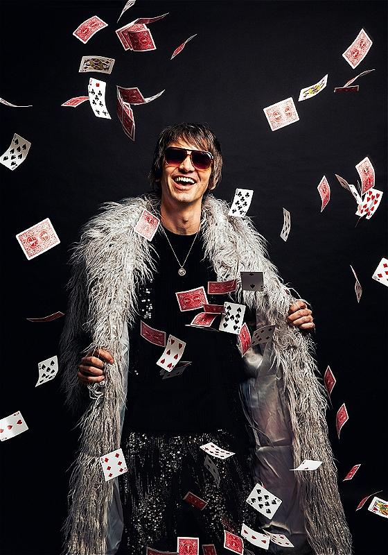 A performer stands against a black background wearing sunglasses and a fur coat. Playing cards are flying down around them.