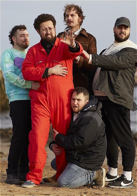 The performers stand on a beach, each with a quizzical expression on their face.