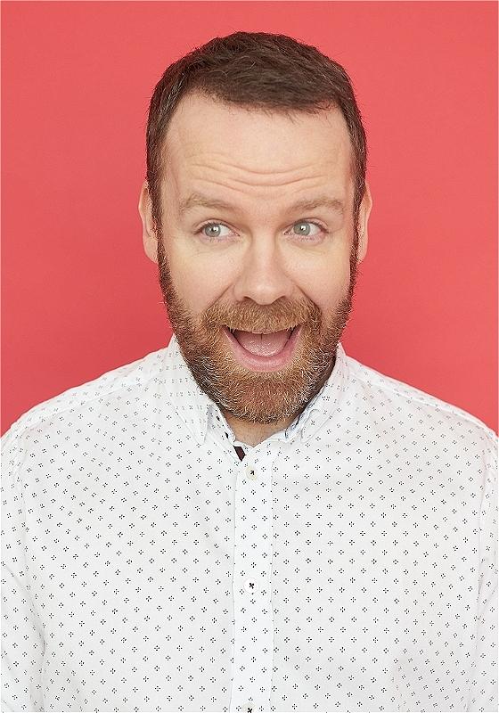 The performer wears a white shirt in front of a red background. His mouth is open and he looks to the side. 