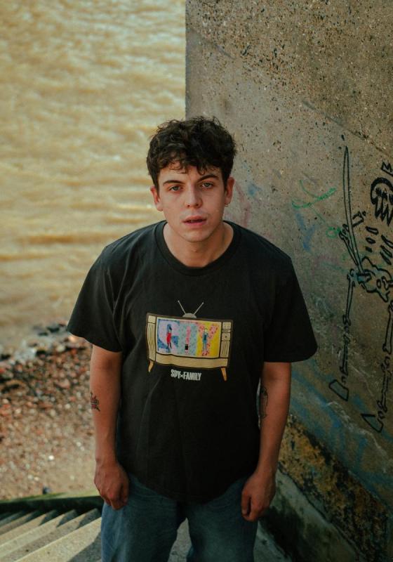 Photo of Ed Night standing by a concrete wall in front of the sea. We see the pebbles on the and Ed standing in front looking up. He wears a black graphic t-shirt.
