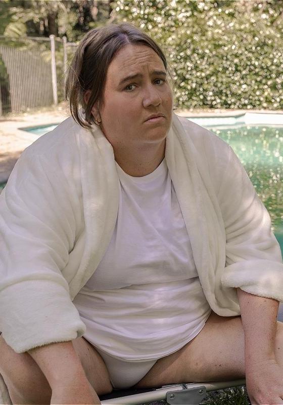 The performer sits in a garden in front of a swimming pool, wearing a white dressing gown.