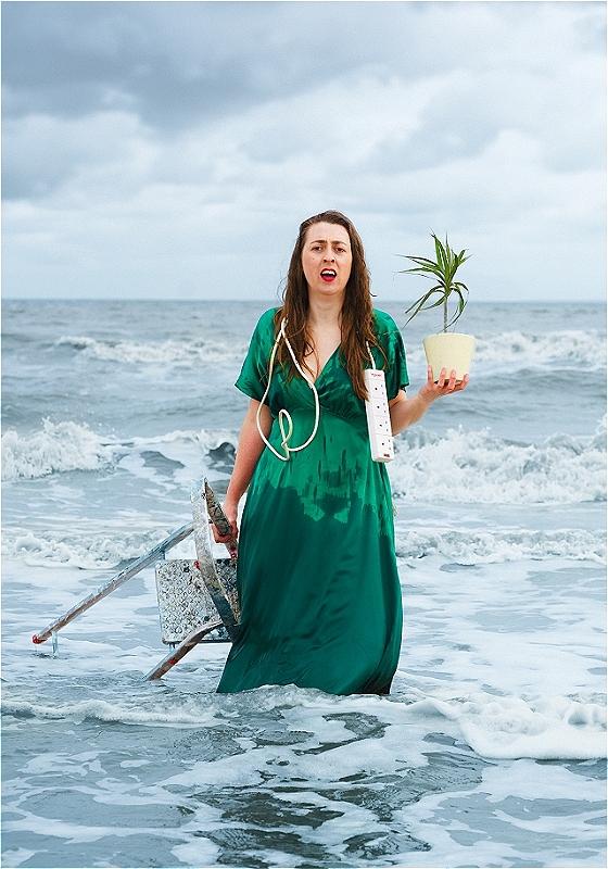 Performer stands in the waves holding a pot plant and ladder with an extension cable draped around their shoulders. The sea spray has left damp splashes all over their green dress.