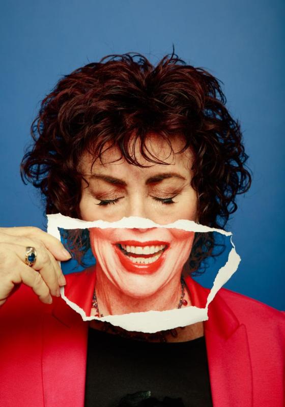 Ruby Wax in front of blue background, holding piece of paper in front of their mouth and smiling through it. 