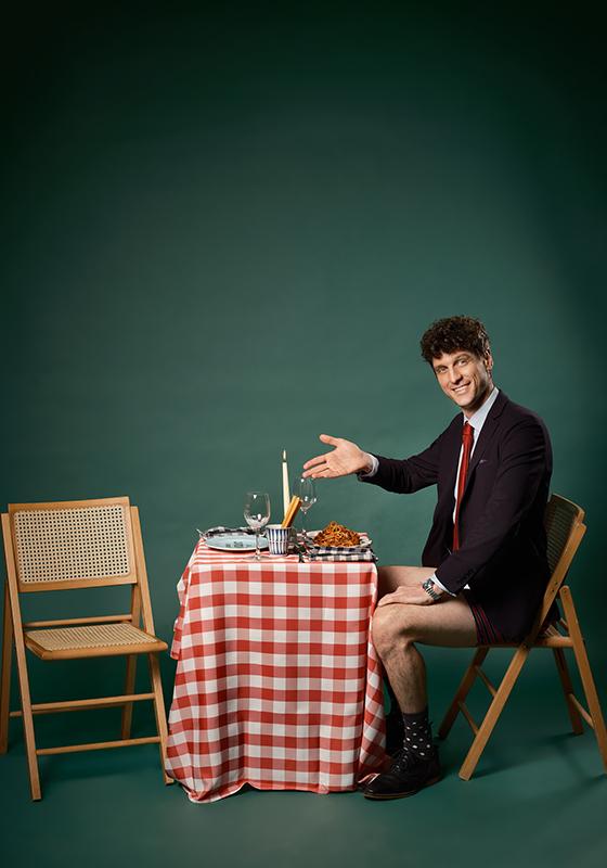 The performer sits at a table with plates and wine glasses. He wears a black suit and underpants, smiling and gesturing to the empty seat opposite. 