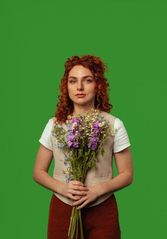 The performer stands in front of a green background. They are holding a bouquet of purple and white flowers.