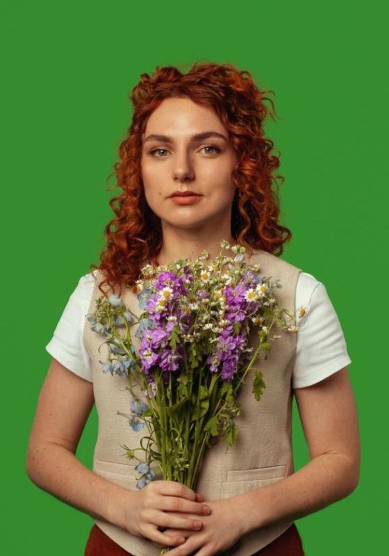 The performer stands in front of a green background. They are holding a bouquet of purple and white flowers.