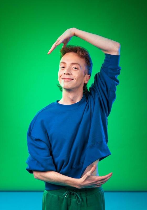 Derek wearing a blue sweatshirt and green trousers, holding one arm above their head. The background is green at the top and blue at the bottom of the image. 