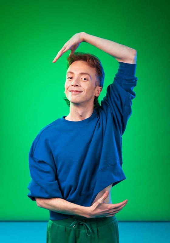 Derek wearing a blue sweatshirt and green trousers, holding one arm above their head. The background is green at the top and blue at the bottom of the image. 