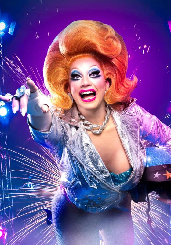 Ginger, in all her glory, wears a fabulous silvery jacket pointing with a hand outstretched into the universe. The lighting is full of blues, pinks and purples and she carries a space helmet with stars in her right hand.