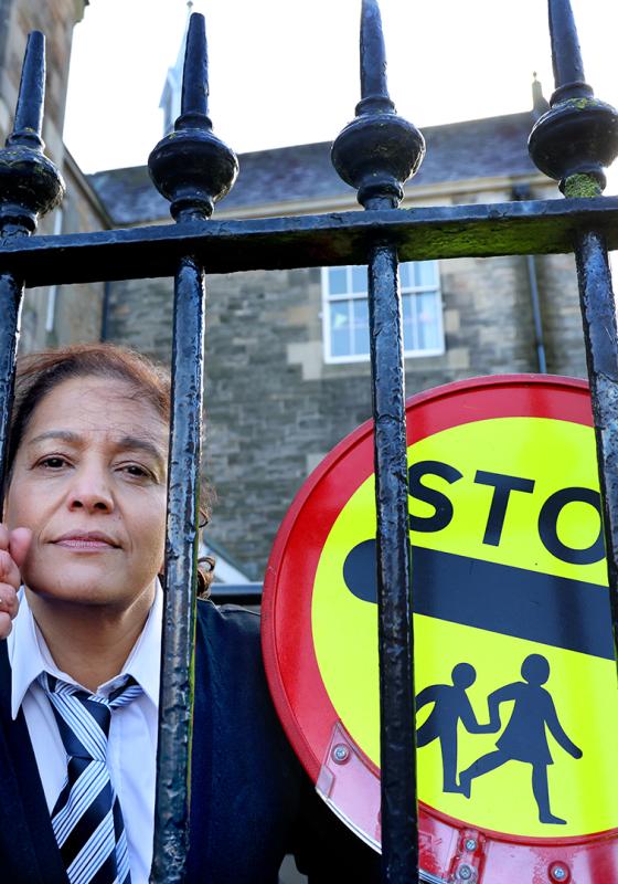 The performer peers through the railing on a school playground, wearing a school uniform and holding a stop sign.