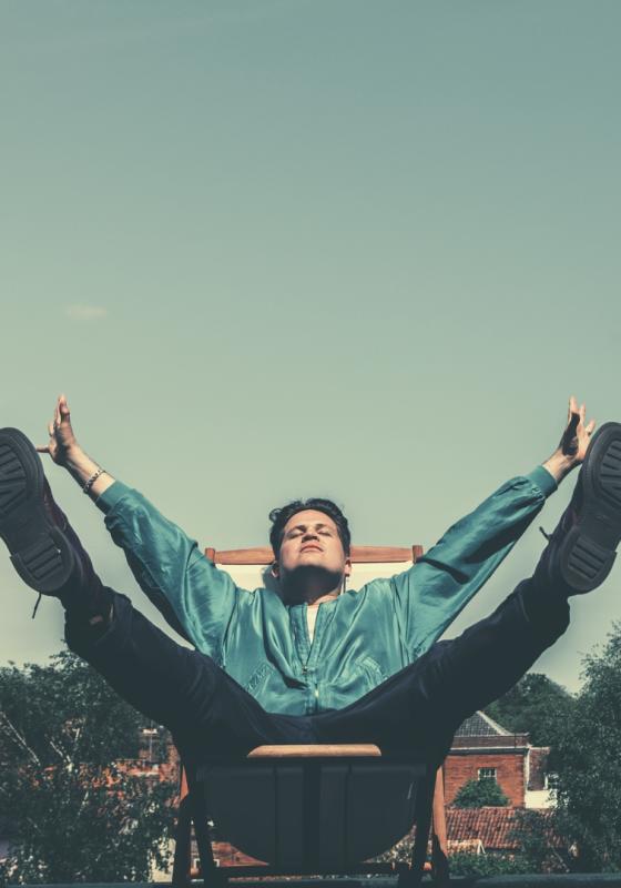 The performer is sat on a deck chair, with their arms and legs spread out reclining in a joyful manner. Houses and trees are in the background.