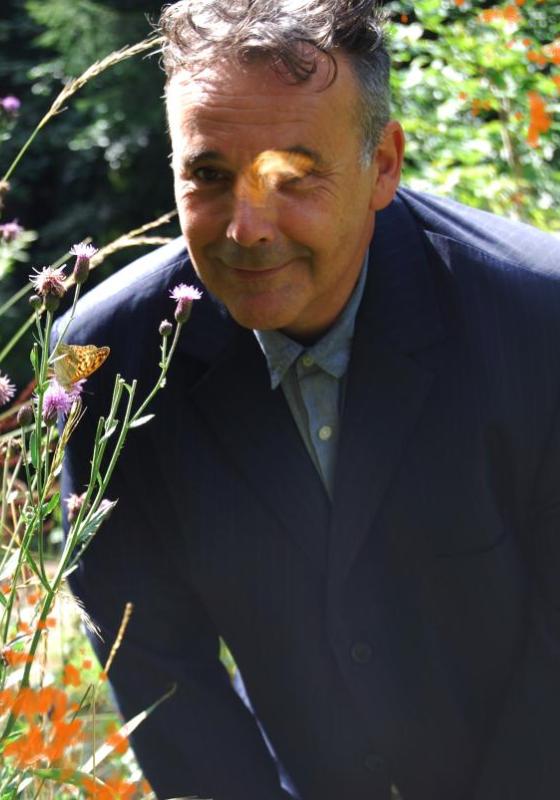 The performer peers through some greenery wearing a suit, smiling at the camera 