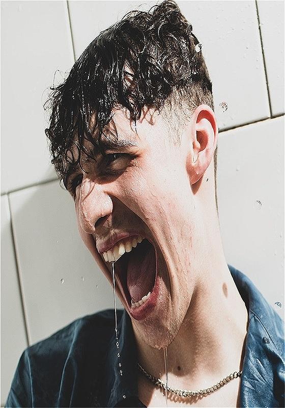 Young man with wet curly hair, wearing a dark shirt, laughing or shouting with joy as water drips from his mouth, against a sunlit white tiled wall.