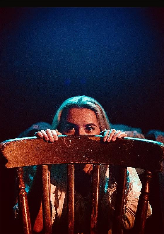 A woman with blonde hair peeking over the top of a wooden chair, her eyes visible as she grips the chair's back. The scene is lit with a warm, theatrical blue light, creating a moody and mysterious atmosphere.