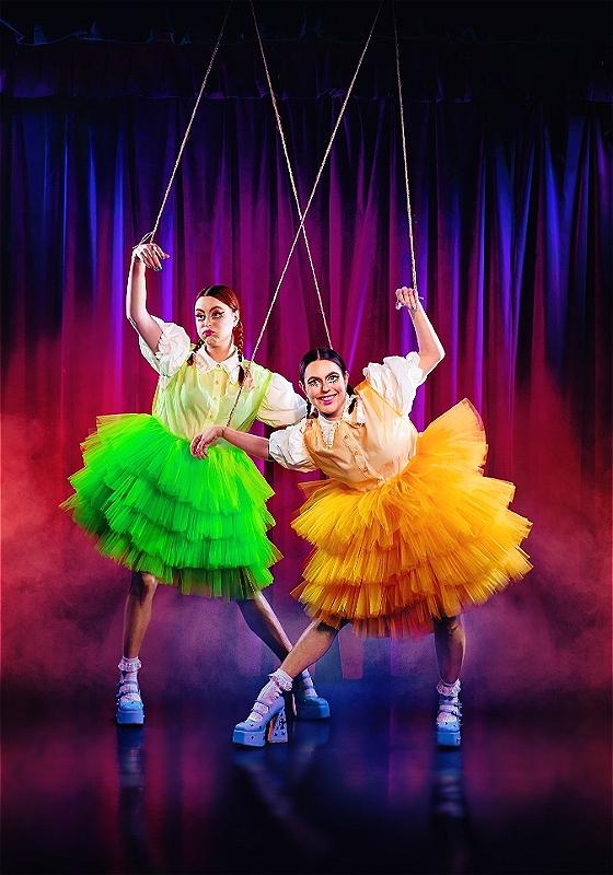 Two performers suspended by strings, resembling marionette puppets, on a stage with a purple curtain background. Both are wearing colorful tutus, the left in green and the right in yellow, with matching suspenders and white socks and high heels. The right performer is smiling at the camera while the left maintains a serious expression.