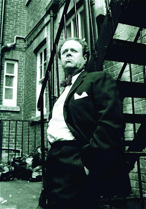 Black and white photo of an older man with receding hair, wearing a suit and polka dot bow tie, standing on a fire escape and looking pensively to the side with a cigarette in his mouth, urban building in the background.