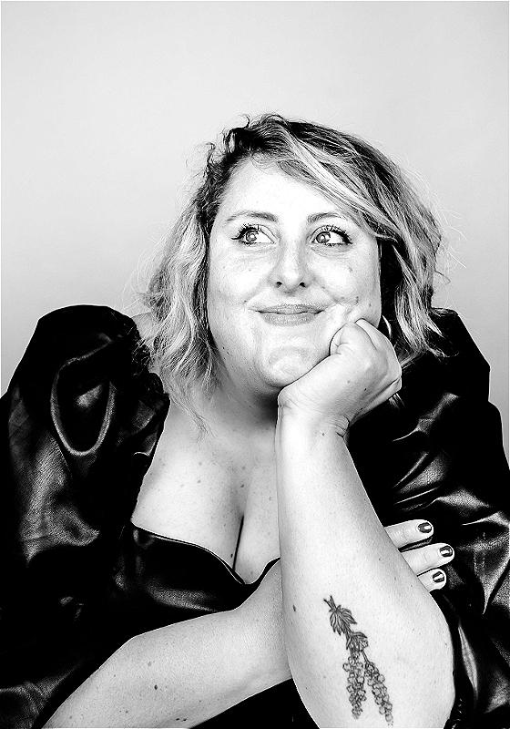 Black and white portrait of a smiling woman with short curly hair, resting her chin on her hand. She wears a black dress with puffy sleeves and shows off a tattoo of grapes on her upper left arm.