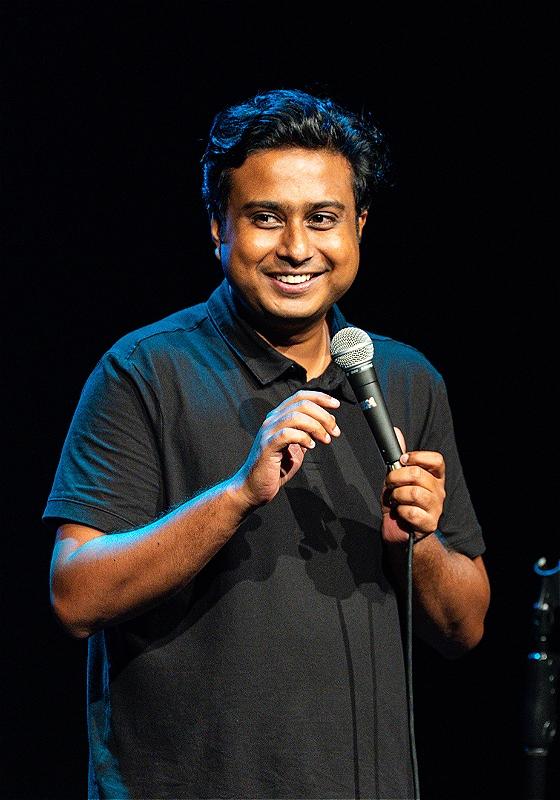 Man standing on stage holding a microphone, smiling slightly during a speech or performance. He is wearing a dark polo shirt with ambient stage lighting highlighting his features.