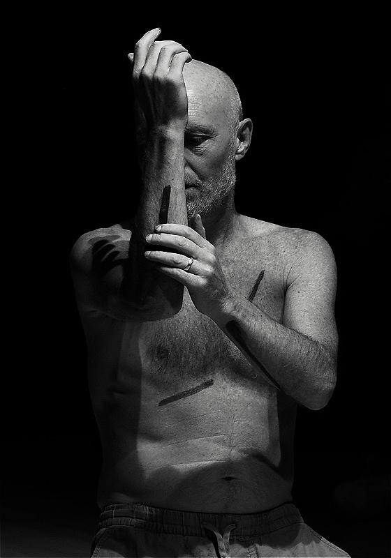 A shirtless performer sits in darkness, with black lines drawn on their body. Their arm is raised in front of their face. 