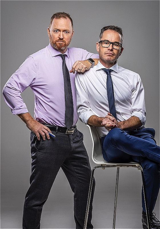 Two men in business attire posing confidently against a gray background; one standing with hands on his hips and the other sitting on a chair with one hand resting on the standing man’s shoulder.