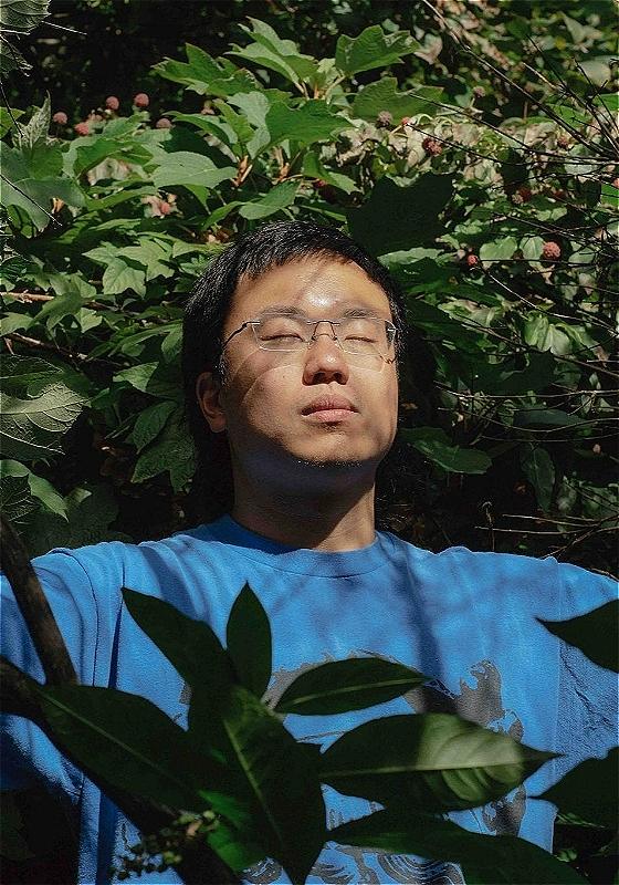 The performer wears a blue tshirt and stands with his eyes closed. He is surrounded by greenery and looks peaceful. 