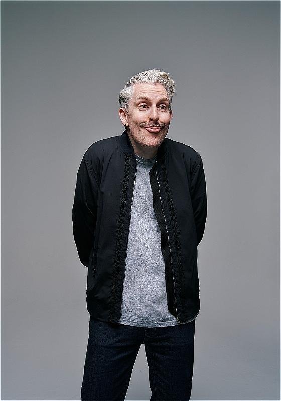 Man with a playful expression and a large, curled mustache standing against a gray background, wearing a black jacket, grey shirt, and dark blue jeans.