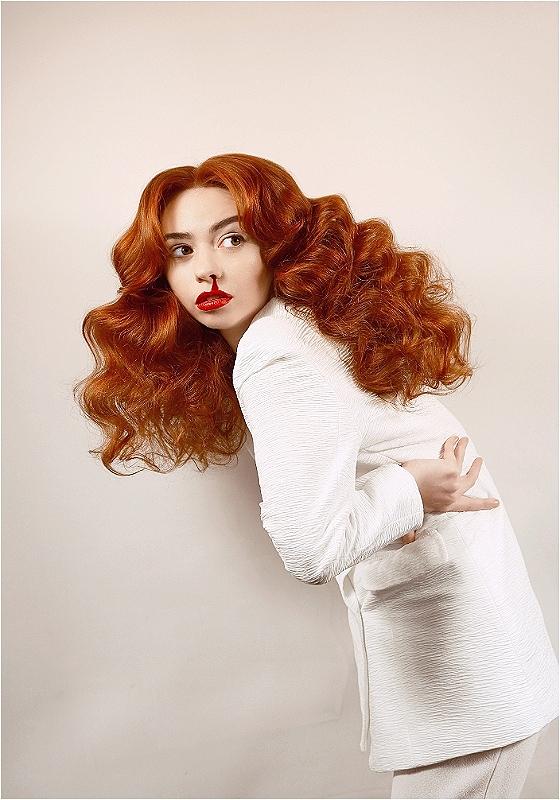 Woman with long curly red hair wearing a white textured dress, standing sideways while turning her head to face the camera with bright red lipstick.