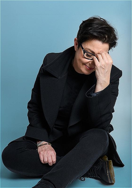 Sue is in black attire sitting, laughing while covering her face with one hand, against a blue background.