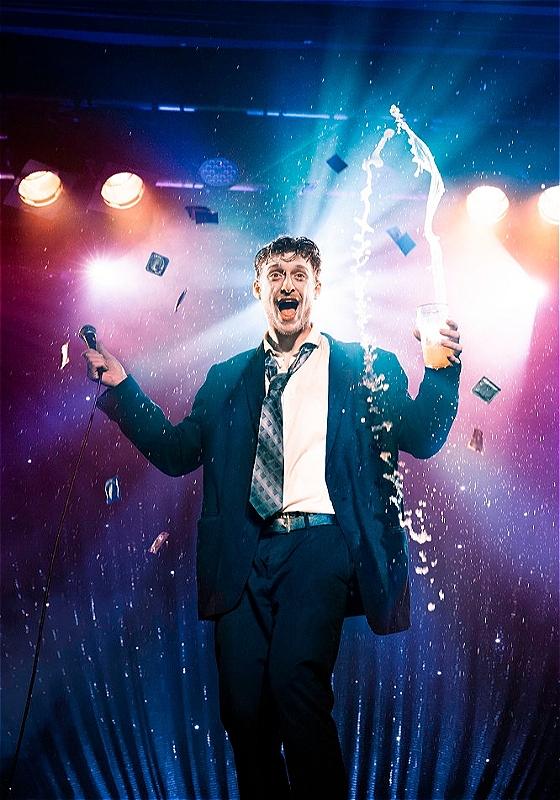 A man enthusiastically singing onstage with a microphone in one hand and a cup spurting liquid in the air in the other, under atmospheric stage lighting with purple and blue tones and floating confetti. He is dressed in a navy suit with a loosened tie, showing an expression of joy and excitement.