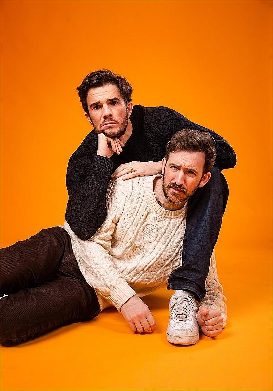 The two performers pose in front of an orange background, with one resting on the other. 