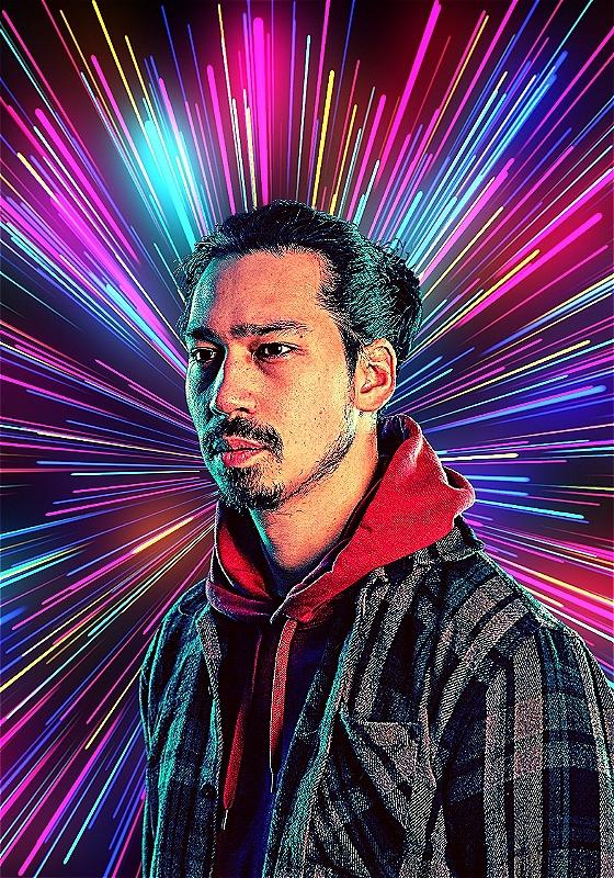 Man with light stubble in a hoodie and jacket, facing slightly left with a serious expression, against a background of vibrant, multicolored light rays emanating from behind him creating a dynamic and futuristic effect.