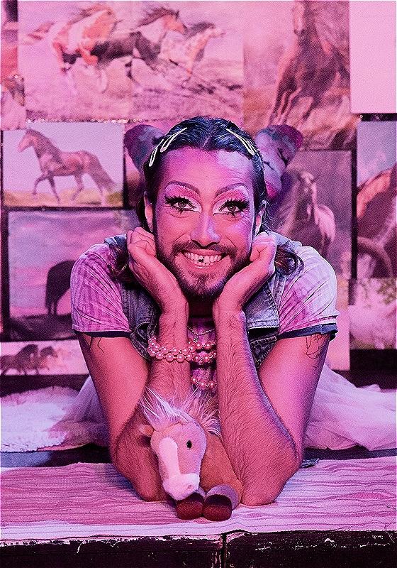 Performer lies on bed smiling at the camera, dressed as a young girl. Pictures of horses cover the walls and a stuffed horse toy is on the centre of the bed.