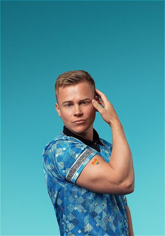 Young man with short blond hair, wearing a blue patterned shirt, poses against a teal background. His right hand is touching his temple, and there is a rainbow heart sticker on his left arm.