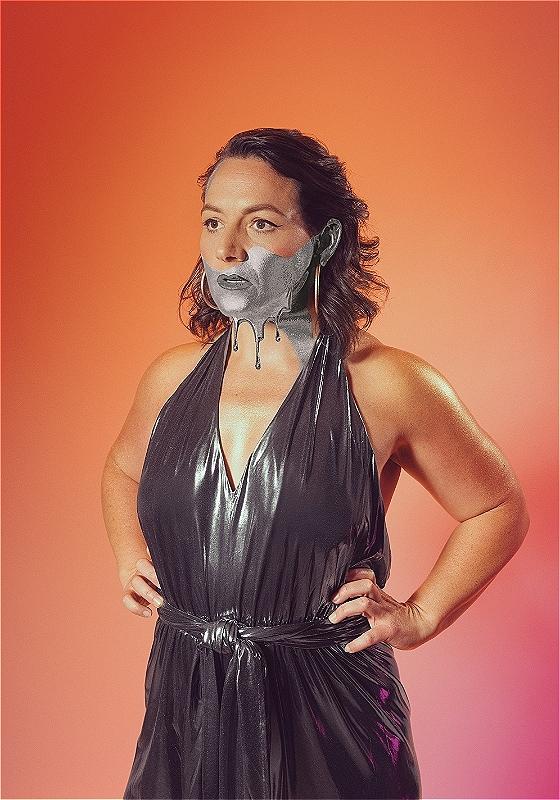 Woman with a metallic silver mask paint on her face, standing against an orange background, wearing a shiny black halter neck dress.